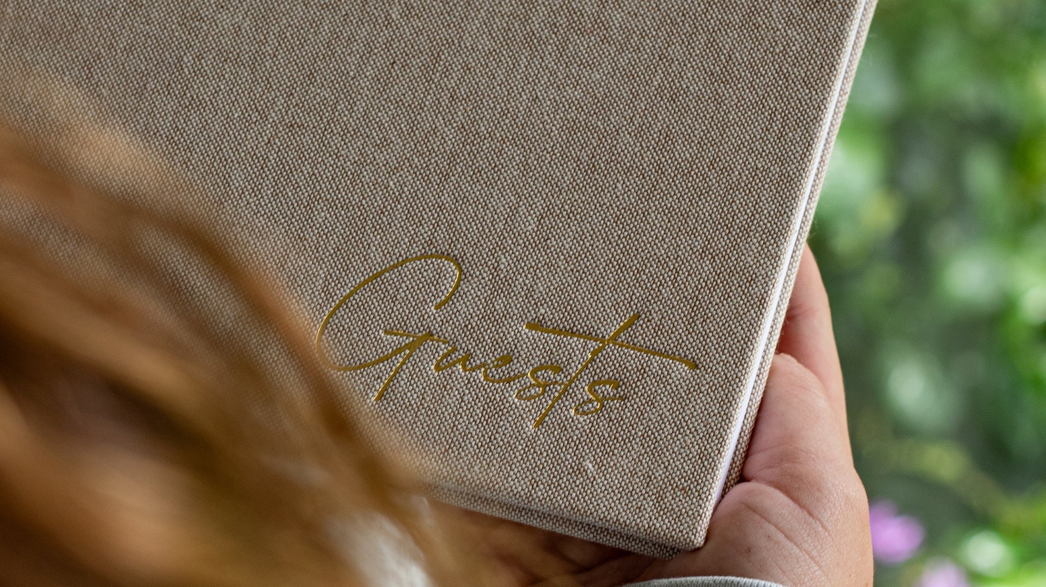 Person holding a linen-covered guestbook with gold foil ‘Guests’ title, standing by a sunny window with greenery outside