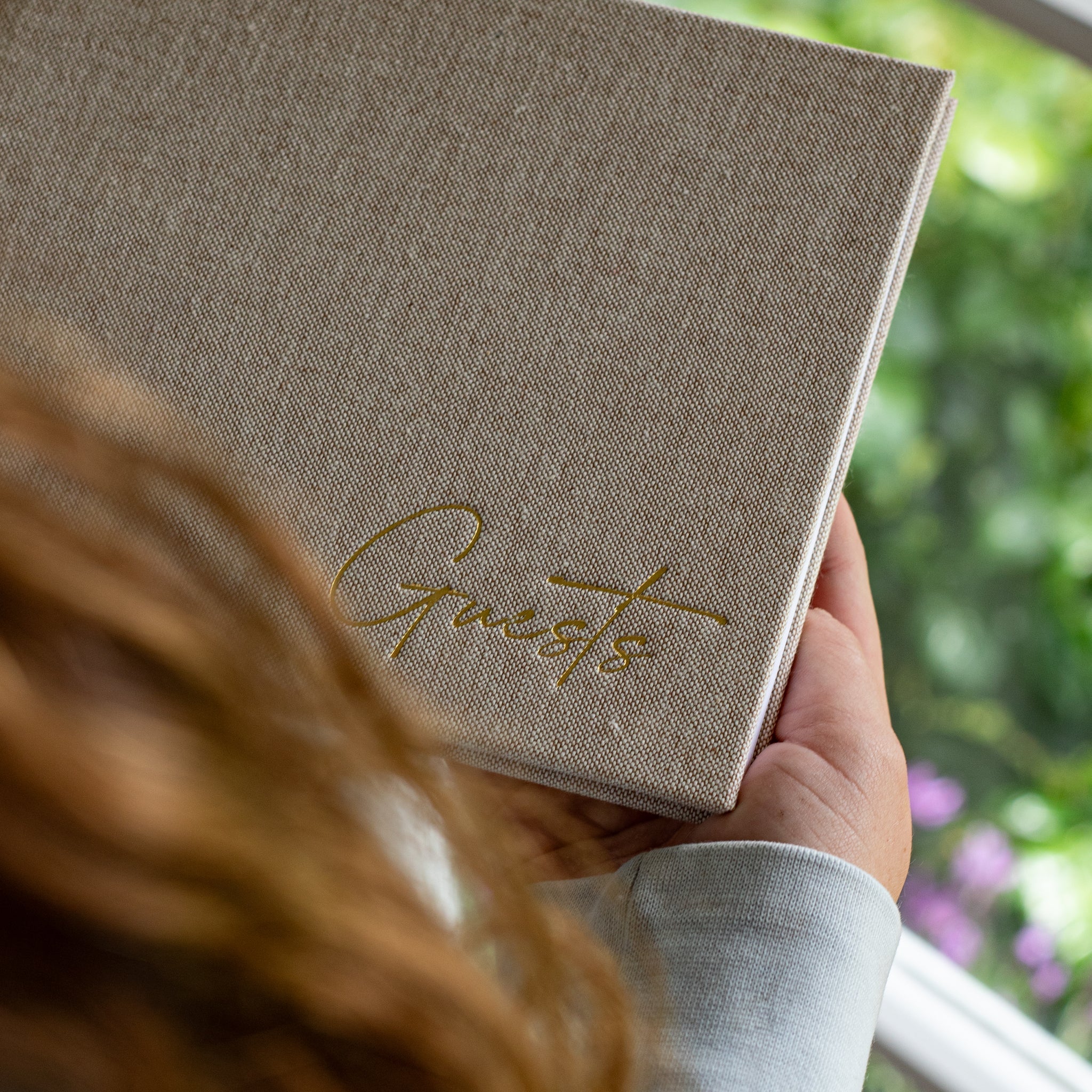 Person holding a linen-covered guestbook with gold foil ‘Guests’ title, standing by a sunny window with greenery outside