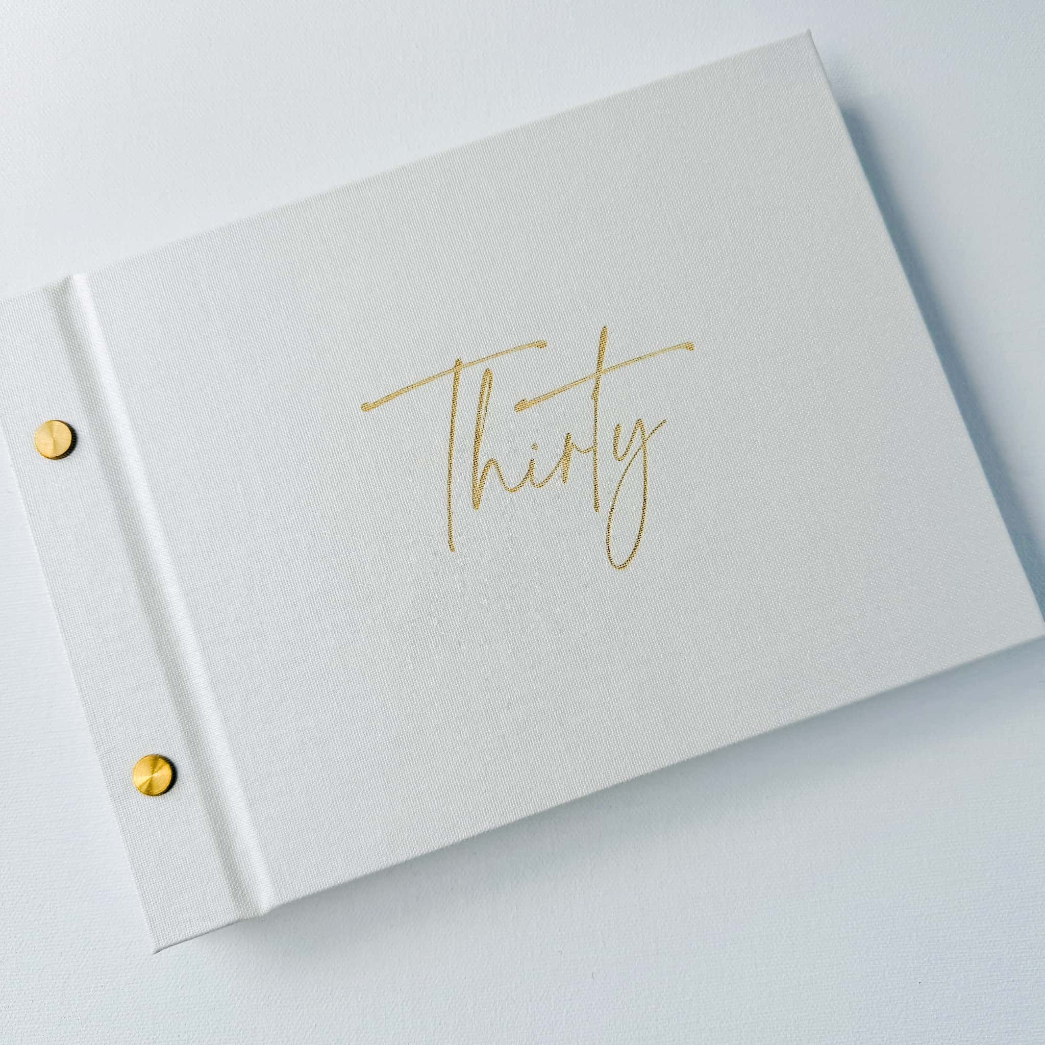 White book with 'Thirty' in gold on a pearl white book cover with gold binding screws