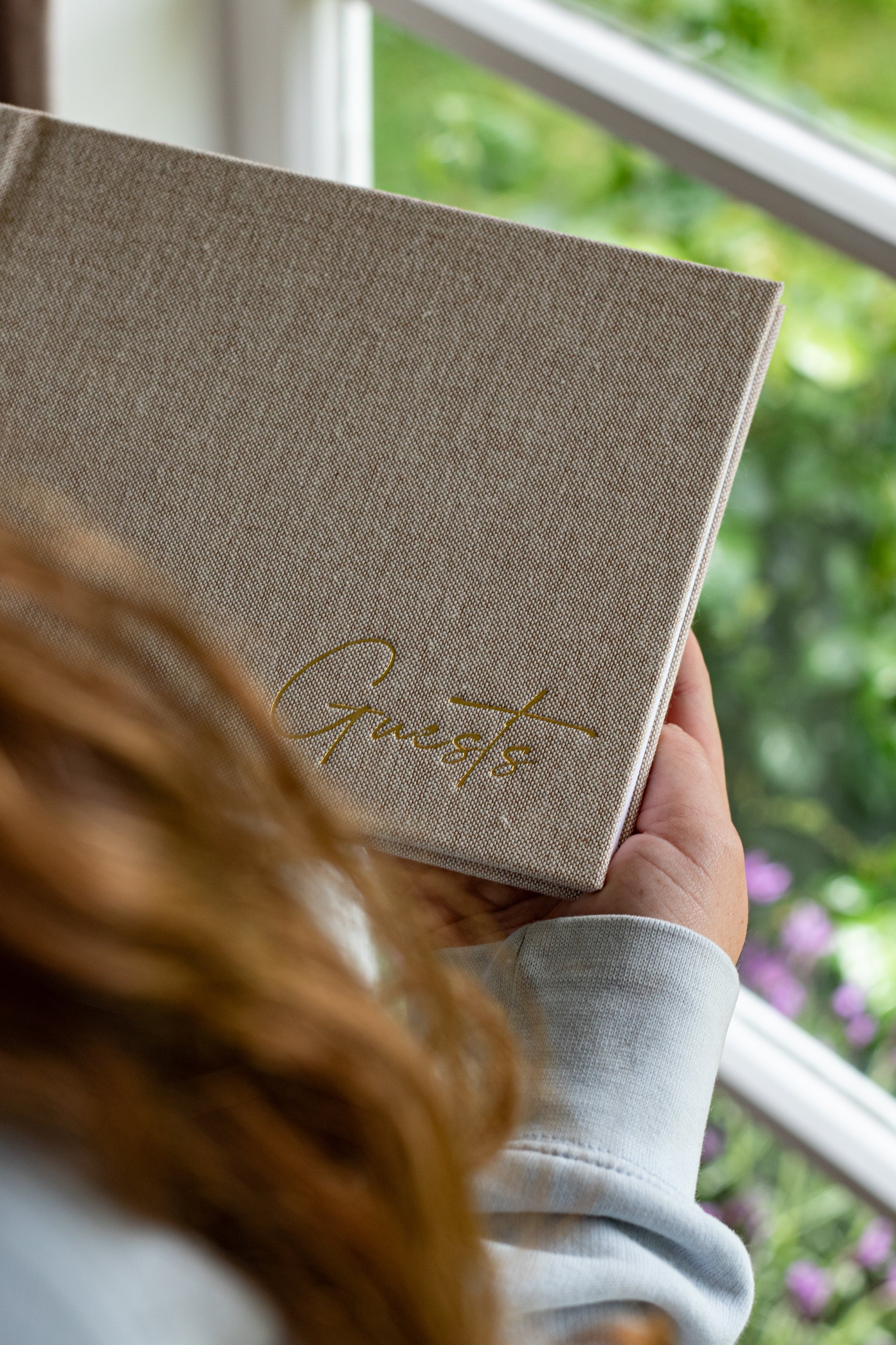 Person holding a linen-covered guestbook with gold foil ‘Guests’ title, standing by a sunny window with greenery outside