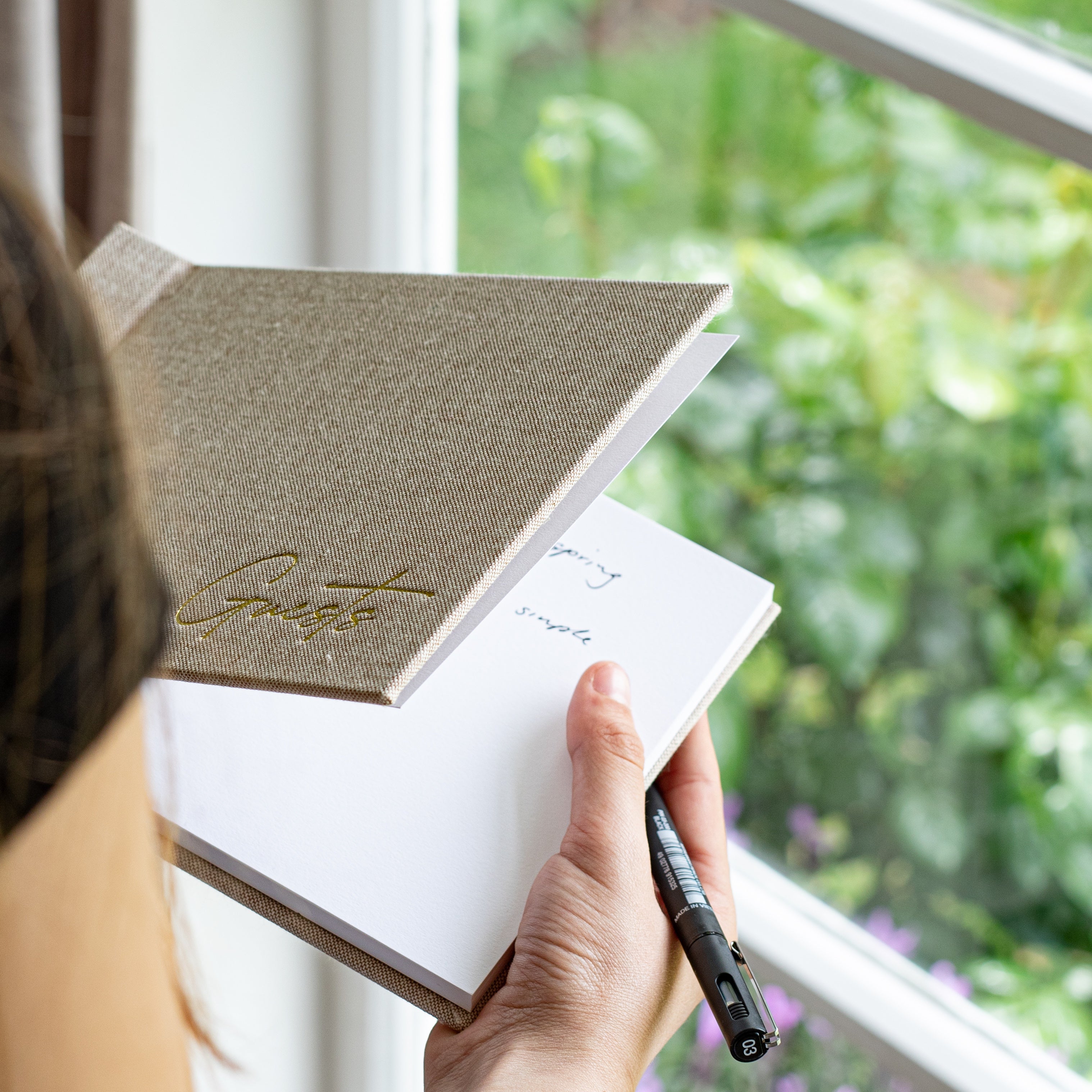 A person holds an open guestbook with a textured oatmeal linen cover and gold script logo, writing in a white notebook with a black pen, near a window with a blurred green garden background.