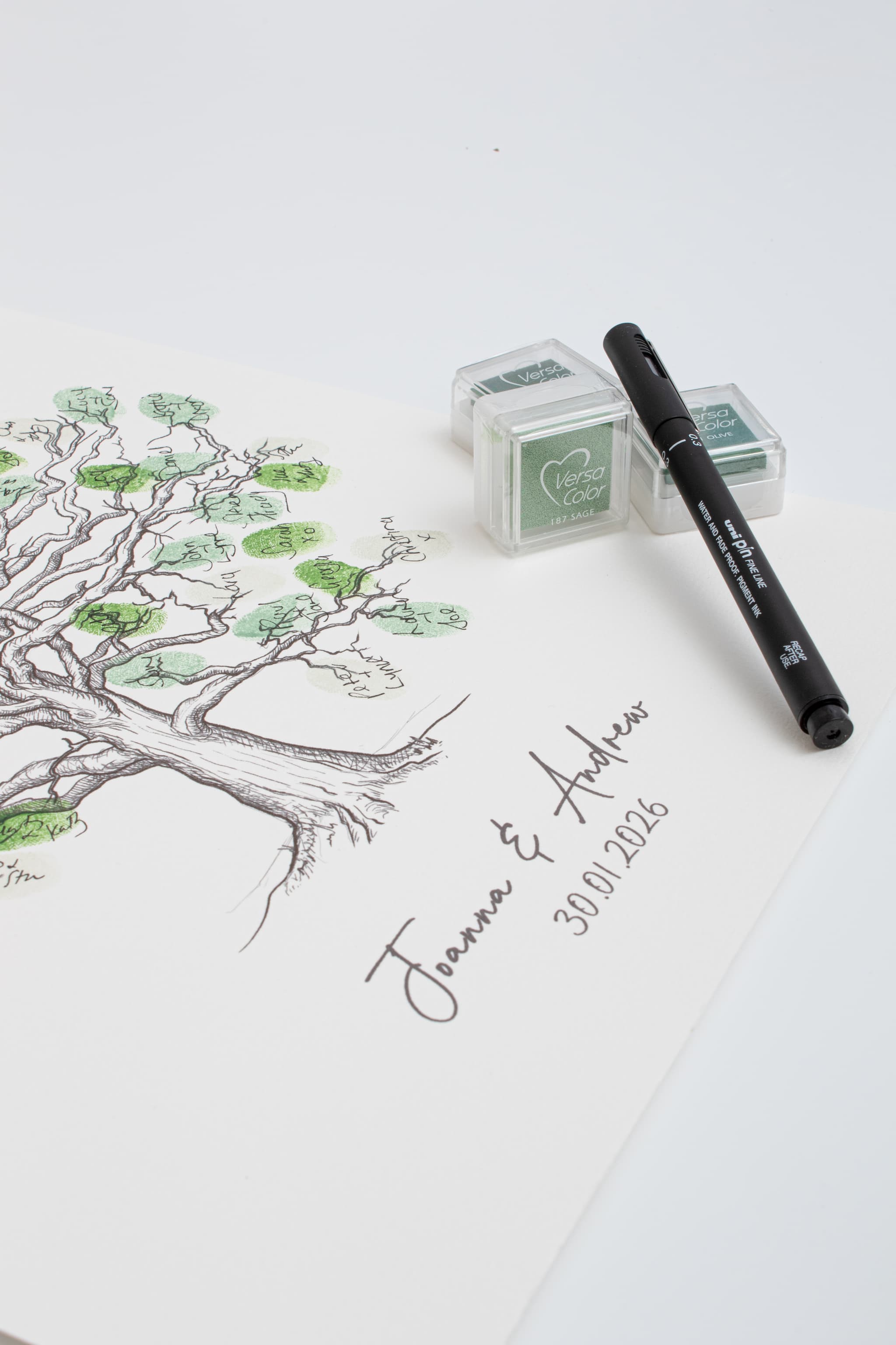 A white paper fingerprint guestbook featuring a black ink oak tree illustration with green leaf accents and the personalised names 'Jenna & Andrew' along with the date '30.01.26'