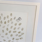 A framed fingerprint guestbook print featuring numerous small, hand-written guest names in muted beige and gold tones, with one larger sketch shaped like a champagne cork. The art piece is housed in a thick, clean white frame.