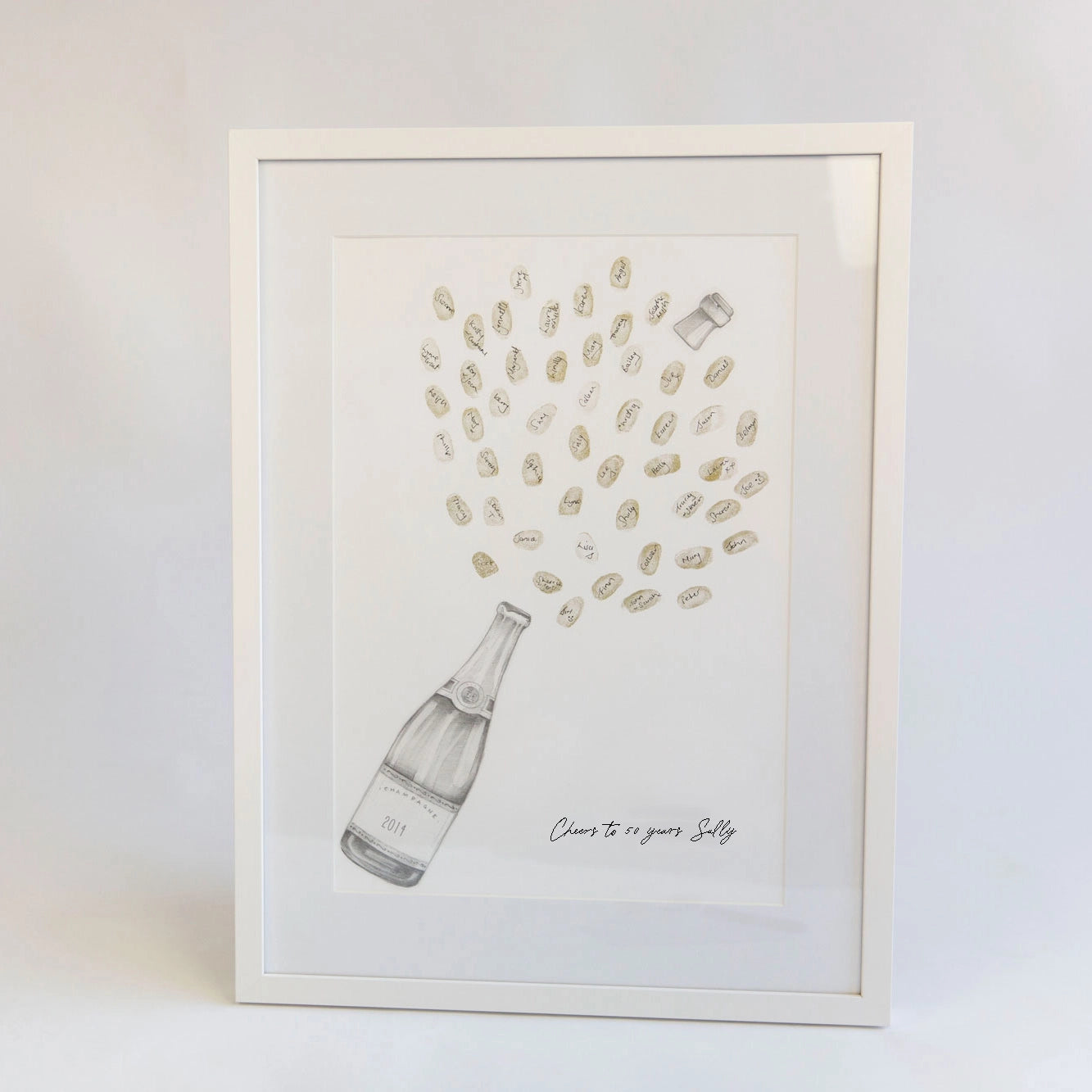 A framed art print featuring a hand-drawn champagne bottle with champagne bubbles bursting out above it, plus the handwritten text 'Cheers to you, Sally'. The print is in a white frame against a clean white background.
