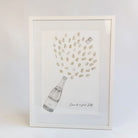 A framed art print featuring a hand-drawn champagne bottle with champagne bubbles bursting out above it, plus the handwritten text 'Cheers to you, Sally'. The print is in a white frame against a clean white background.