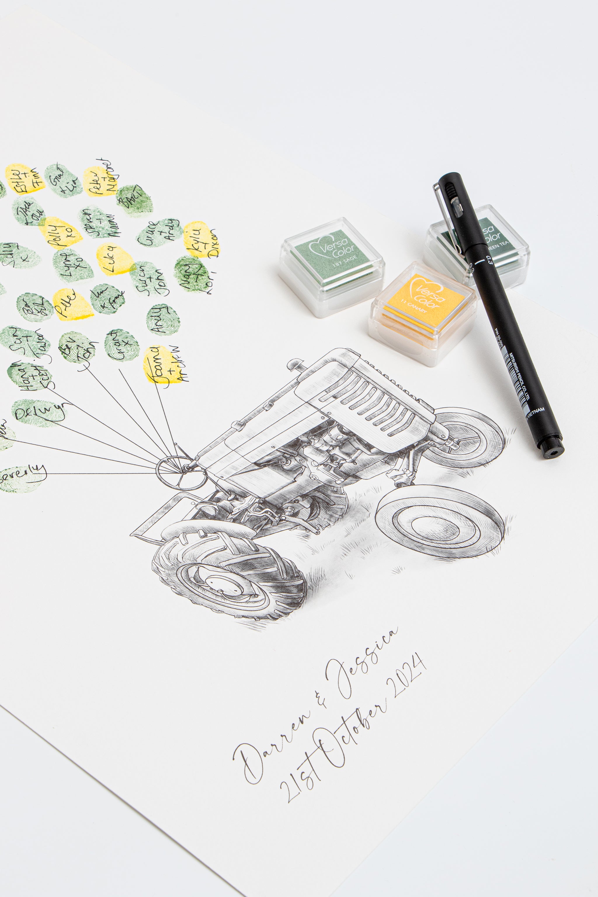 A custom fingerprint wedding guestbook with a vintage tractor design printed on white paper, styled with green and yellow ink stamps, a black pen, and two ink pads on a clean white background.