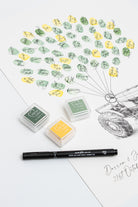 Three small ink pads in green and yellow shades, a black pen, and a printed fingerprint wedding guestbook with many stamped guest fingerprints and signatures arranged like a balloon lifting the tractor off the ground photographed on a white background.