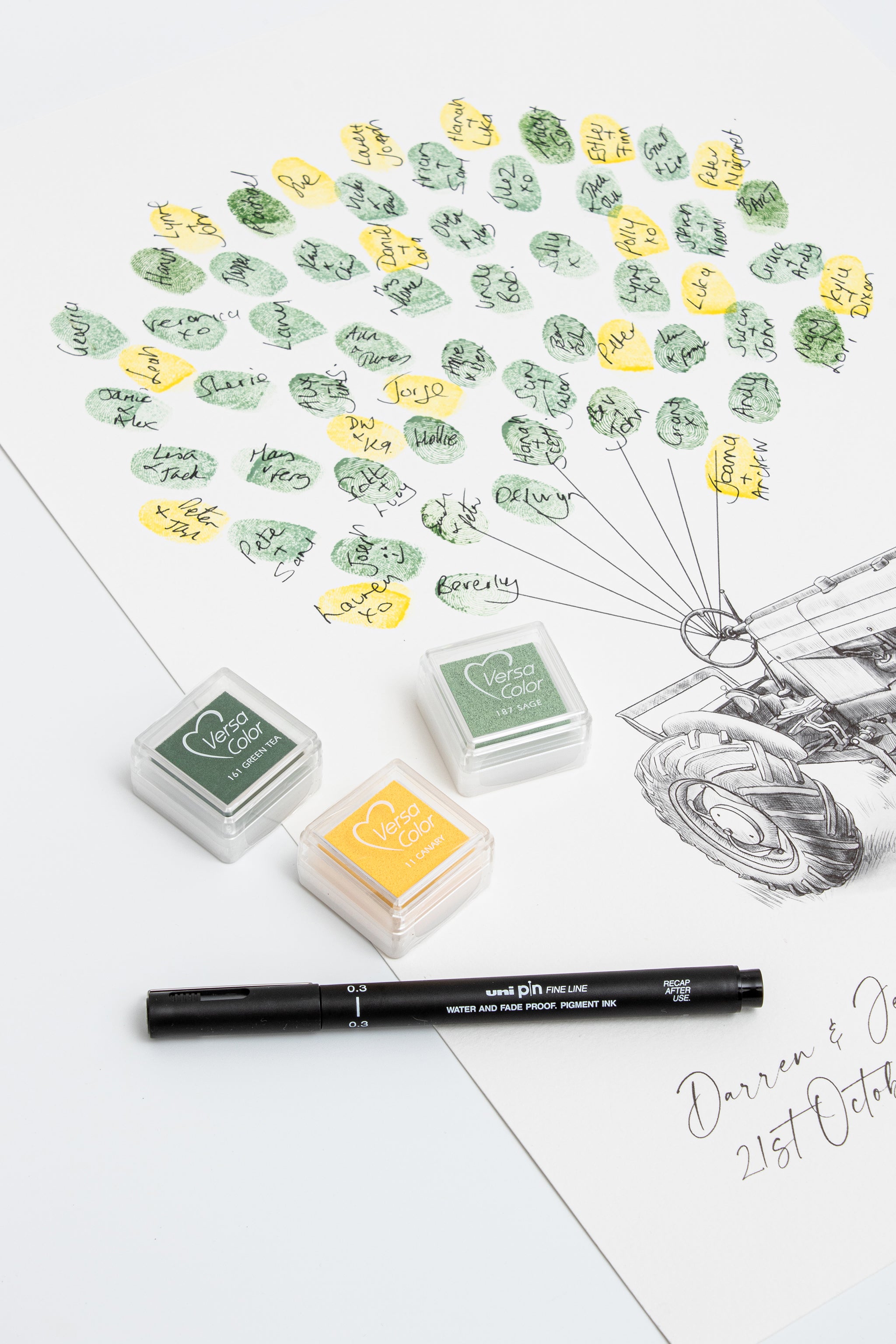Three small ink pads in green and yellow shades, a black pen, and a printed fingerprint wedding guestbook with many stamped guest fingerprints and signatures arranged like a balloon lifting the tractor off the ground photographed on a white background.